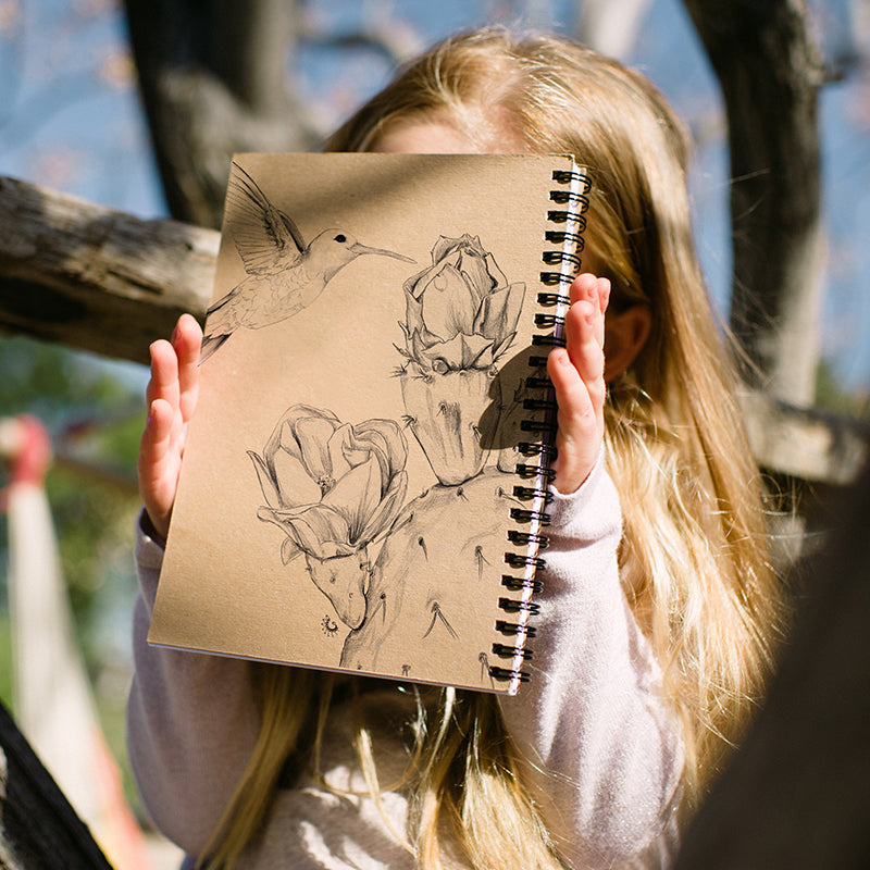 ילדה מחזיקה מחברת שורות A5 בכריכת ספירלה עם רישום עיפרון של יונק דבש ליד קקטוס.